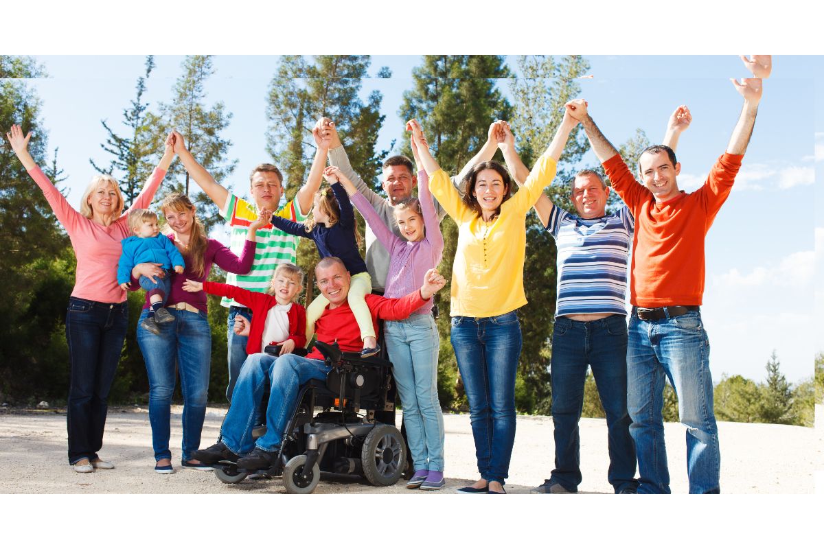 Group of families outdoors raising joined hands, including a man using a power wheelchair, smiling in front of trees