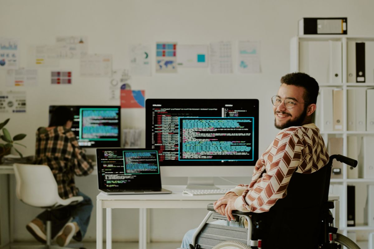 Developer using a wheelchair working at a desk with multiple monitors showing code, smiling at the camera